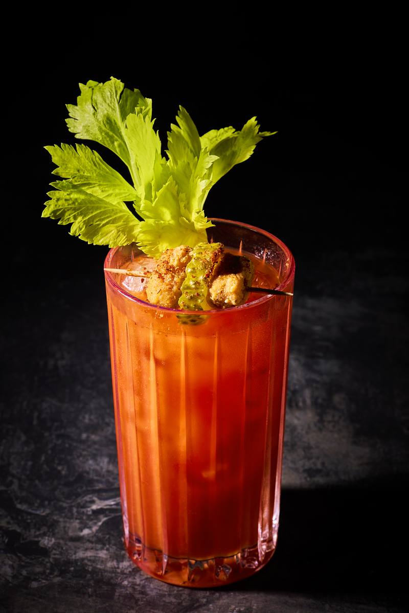 A Bloody Mary in a ribbed glass with celery leaves and a crispy popcorn chicken garnish on a pick